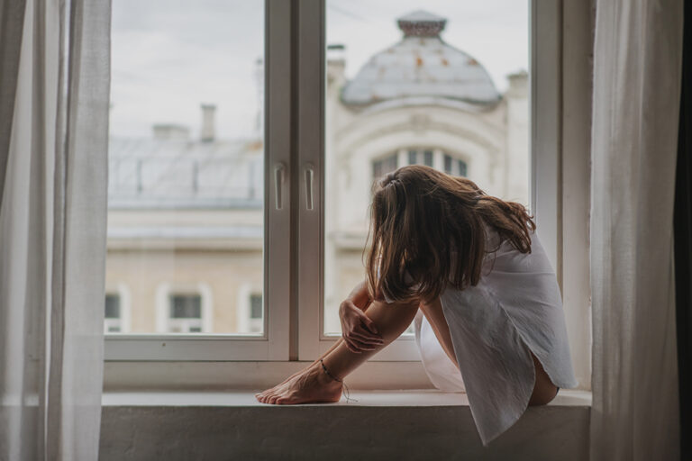 Young sad woman sitting on the windowsill by the window in the a